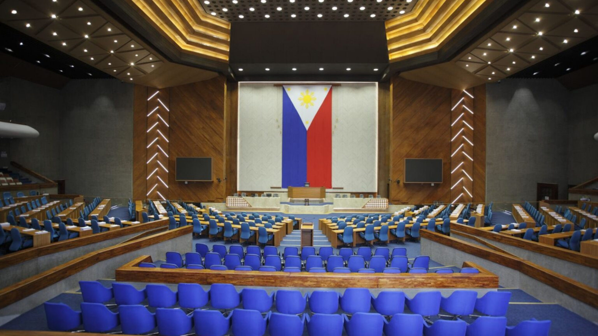 The empty plenary hall of the Philippine House of Representatives in a 2023 photograph, with blue seating facing the rostrum and a large Philippine flag.